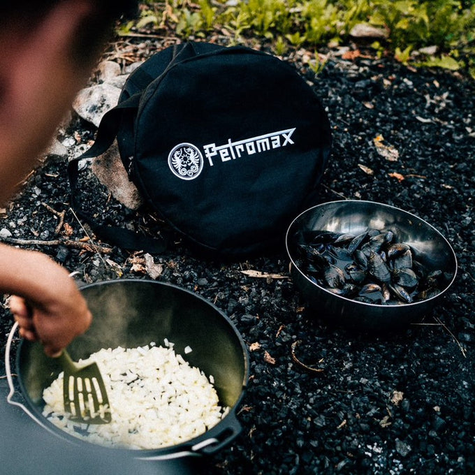Black round carrying case with Petromax logo, designed for storing and transporting Dutch ovens, shown outdoors beside cooking pots with food, suited for camping and outdoor use in farm and garden collections.