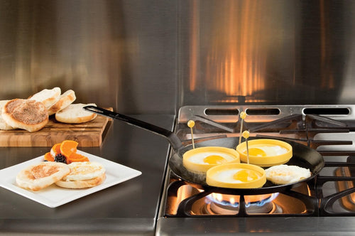 Black seasoned steel skillet with long handle cooking eggs in three yellow silicone egg rings on a gas stove burner, accompanied by toasted English muffins on a white plate and wooden cutting board, perfect for Kitchen & Food, Cookware, Pots and Pans, USA Made, Best Sellers, and Promo Eligible collections.