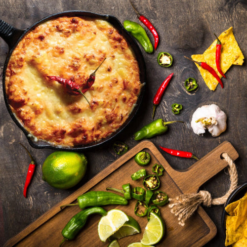Baked cheesy dip in a black cast iron skillet topped with a dried chili, surrounded by fresh jalapeños, garlic, lime wedges, chili peppers, and tortilla chips, perfect for Kitchen & Food, Dry Goods and Mixes, Best Sellers, Promo Eligible collections.