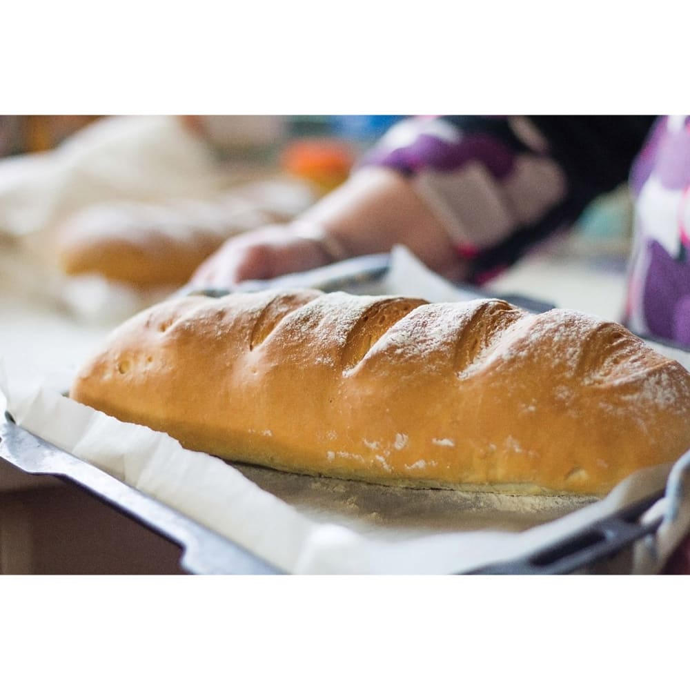 Freshly baked artisan bread loaf resting on parchment paper in a baking pan, showcasing a golden crust with scored slashes, ideal for Kitchen & Food and Bread Making Supplies collections.