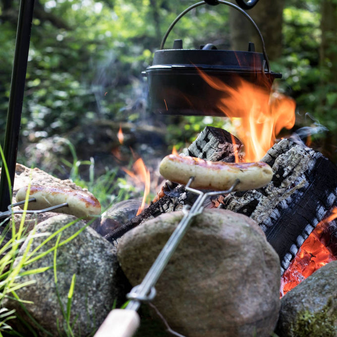Handcrafted campfire skewer with adjustable handle roasting sausage over open flame, surrounded by natural stones, ideal for Farm, Garden & Tool, Camping, Best Sellers, and Promo Eligible collections.