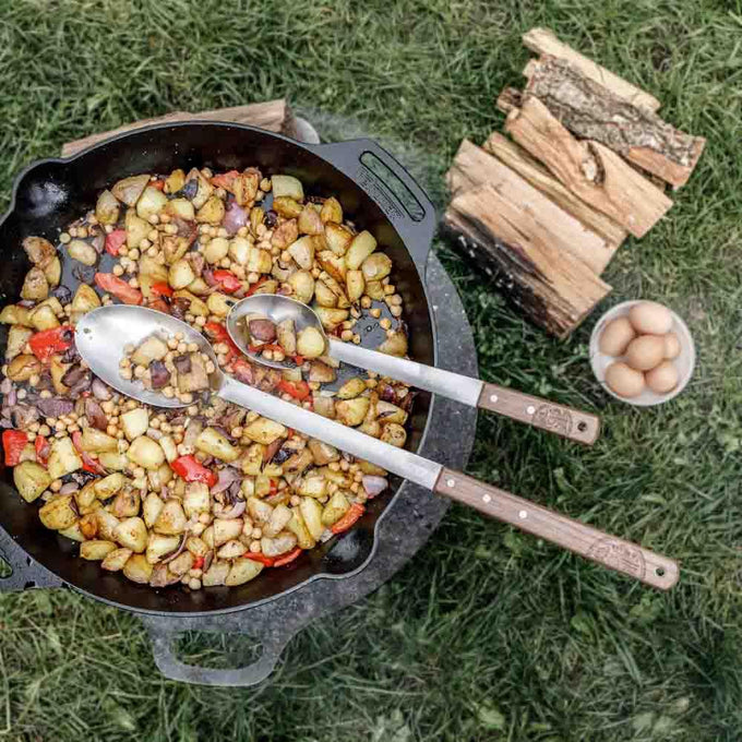 Two handcrafted long serving spoons with stainless steel bowls and walnut handles resting in a cast iron skillet filled with cooked vegetables and chickpeas, outdoors on grass near firewood and a bowl of eggs, ideal for Farm, Garden & Tool, Kitchen & Food, Camping, Storage and Serving, Best Sellers, Promo Eligible collections.