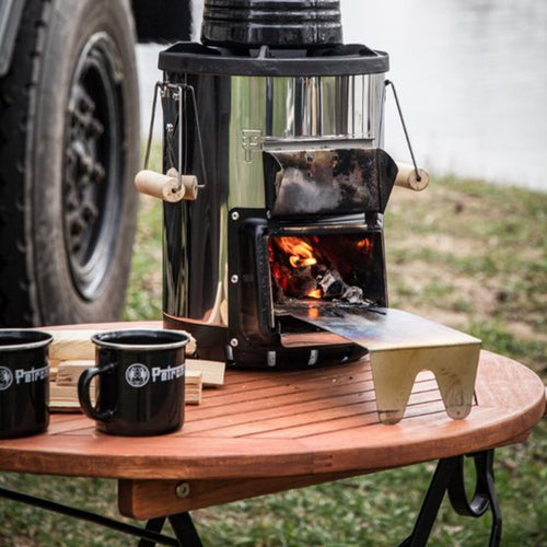 Portable wood stove with polished steel body and wooden handles burning firewood on a round wooden table outdoors, accompanied by two black enamel camping mugs, suited for Farm, Garden & Tool, Kitchen & Food, Camping, Brewing Coffee and Tea, Dinnerware, and Best Sellers collections.
