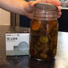 Hand placing a wide-mouth canning lid on a glass jar filled with pickles, next to a box of 12 BPA-free lids, part of Kitchen & Food, Food Preservation, Canning, and Best Sellers collections.