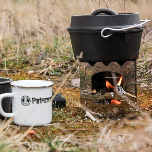 Black cast iron Dutch oven with legs heating over a compact outdoor wood-burning stove, set on grassy ground alongside a white enamel mug, ideal for Kitchen & Food, Cookware, Cast Iron, Best Sellers, and Promo Eligible collections.