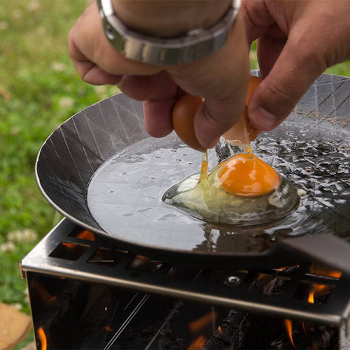 Handcrafted wrought iron pan with a textured cooking surface heating over a portable outdoor stove as an egg is cracked into the pan, ideal for Farm, Garden & Tool, Kitchen & Food, Camping, Cookware, Pots and Pans, Best Sellers, and Promo Eligible collections.