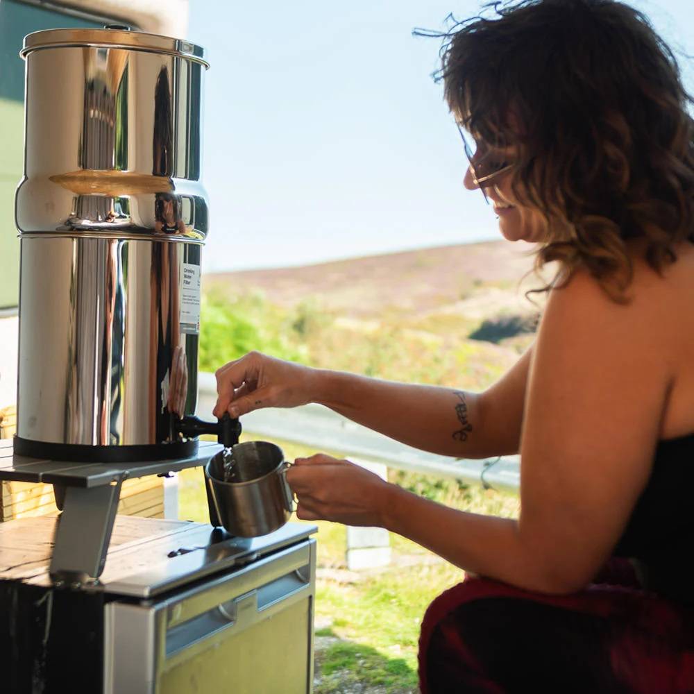 Woman using a handcrafted stainless steel gravity water filter to fill a cup outdoors, ideal for camping, emergency supplies, and farm, garden & tool use.