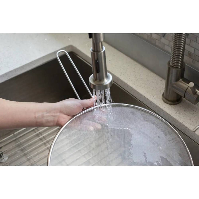 Handheld stainless steel splatter screen being rinsed under running faucet in kitchen sink, designed to protect countertops while cooking, ideal for Kitchen & Food and Cooking Helpers collections.