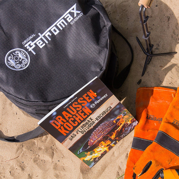 Black durable transport bag with sturdy straps, shown alongside orange heat-resistant gloves and an outdoor cooking book, ideal for camping and farm tool collections.