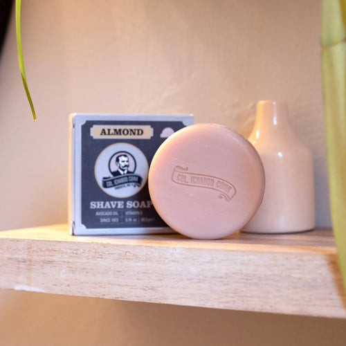 Round almond-scented shaving soap bar with embossed logo, displayed next to its boxed packaging on a wooden shelf, featured in Health & Wellness and Grooming collections.