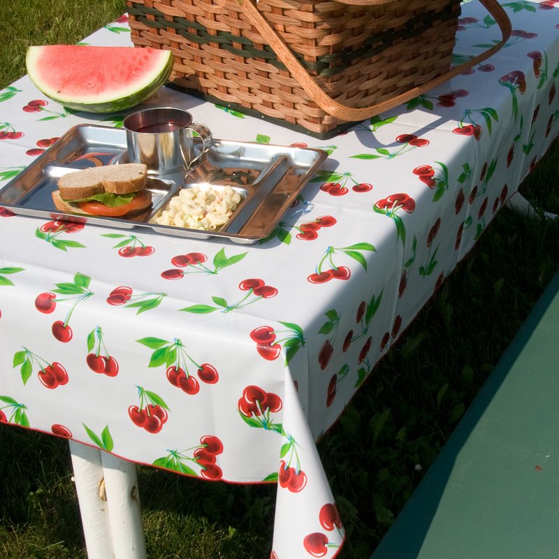 Rectangular vintage vinyl tablecloth featuring a bright cherry pattern with red merrowed edges, displayed outdoors with a picnic basket and lunch tray, ideal for home and kitchen textiles.
