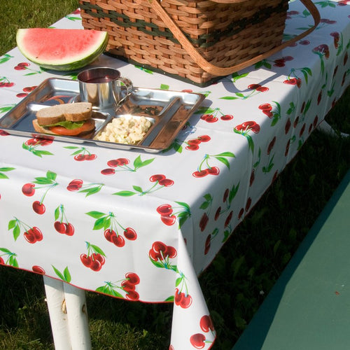 Rectangular vintage vinyl tablecloth featuring a bright cherry pattern with red merrowed edges, displayed outdoors with a picnic basket and lunch tray, ideal for home and kitchen textiles.