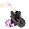 Handheld wooden stomper pressing into a dark ceramic crock with lid resting beside it, surrounded by fresh purple cabbage, for kitchen, food preservation, and fermenting use.