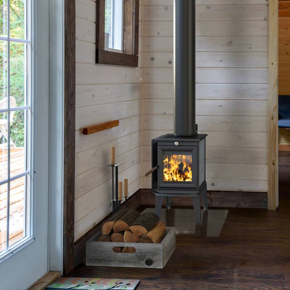 Compact black metal wood heat stove with roaring fire visible through glass door, fitted with a vertical flue pipe, situated in a cozy cabin corner with stacked firewood nearby, ideal for stoves, heatstoves, and best sellers collections.