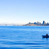 Calm blue waters with a lone person rowing a small boat toward a distant city skyline under a clear sky, evoking tranquility for Best Sellers and P3 Testing collections.