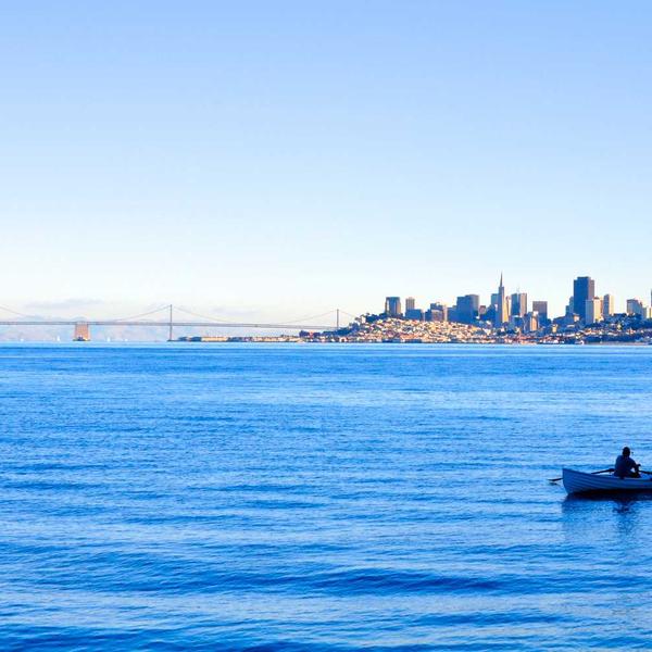 Calm blue waters with a lone person rowing a small boat toward a distant city skyline under a clear sky, evoking tranquility for Best Sellers and P3 Testing collections.