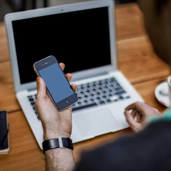 Person holding a smartphone with a blank screen in front of a laptop on a wooden table, suggesting remote work or digital testing related to Best Sellers and P3 Testing collections.