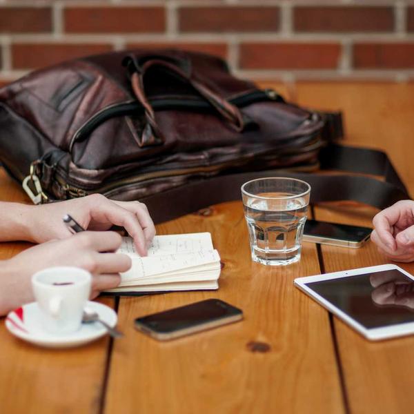 Two people engaged in discussion at a wooden table featuring a leather bag, notebook, smartphone, tablet, glass of water, and espresso cup, evoking a focused and productive moment for Best Sellers and P3 Testing collections.