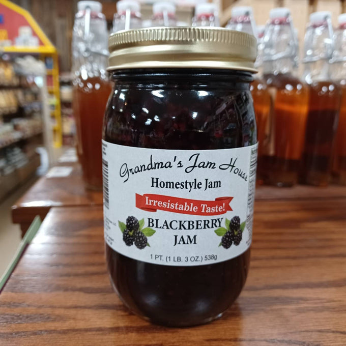 Jar of blackberry jam with gold lid and label reading "Grandma's Jam House Homestyle Jam" on wooden table, surrounded by glass bottles, featured in Kitchen & Food, Food, Pantry, Peanut Butter & Jellies, USA Made, Best Sellers, and Promo Eligible collections.