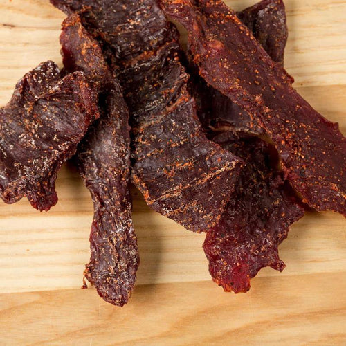 Close-up of several dark, seasoned jerky strips laid on a light wooden surface, showcasing a gluten-free, handcrafted, protein-rich snack made in USA for Kitchen & Food, Meats, and Best Sellers collections.