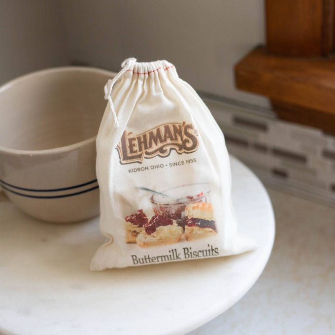 Soft cloth drawstring bag of handcrafted Lehman's buttermilk biscuit mix with image of golden biscuits, sitting on a kitchen table near a pottery bowl, ideal for Kitchen & Food, Pantry, and Dry Goods collections.