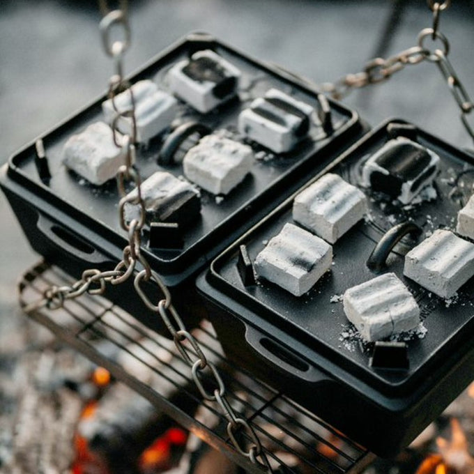 Two cast iron loaf pans filled with charcoal briquettes cooking over an open campfire grill, showcasing durable cookware designed for farm, kitchen, camping, and bread making supplies collections.