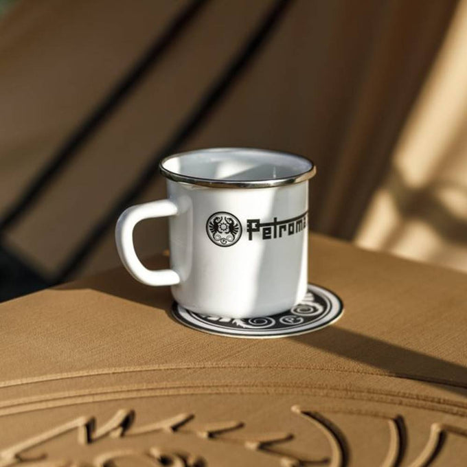 White enameled steel mug with black logo and metal rim, sitting on a textured surface in natural light, ideal for Farm, Garden & Tool, Camping, Kitchen & Food, and Brewing Coffee and Tea collections.