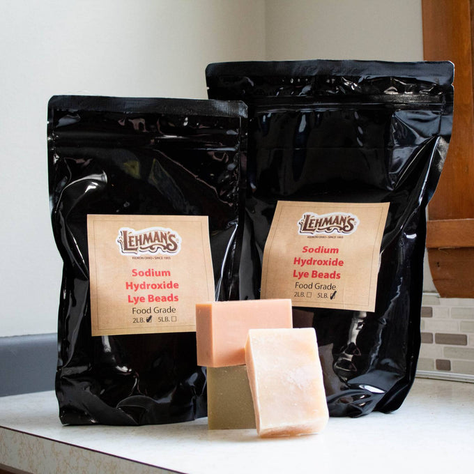 Two resealable black bags of food-grade sodium hydroxide lye beads in 2-lb and 5-lb sizes with handcrafted soap bars in front, displayed in a kitchen setting.