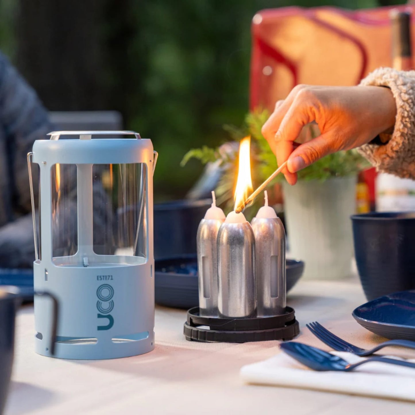 Handcrafted candle lantern with a light gray metal frame beside a set of three tall replacement candles being lit outdoors, suited for camping, emergency, and outdoor use in farm and garden settings.