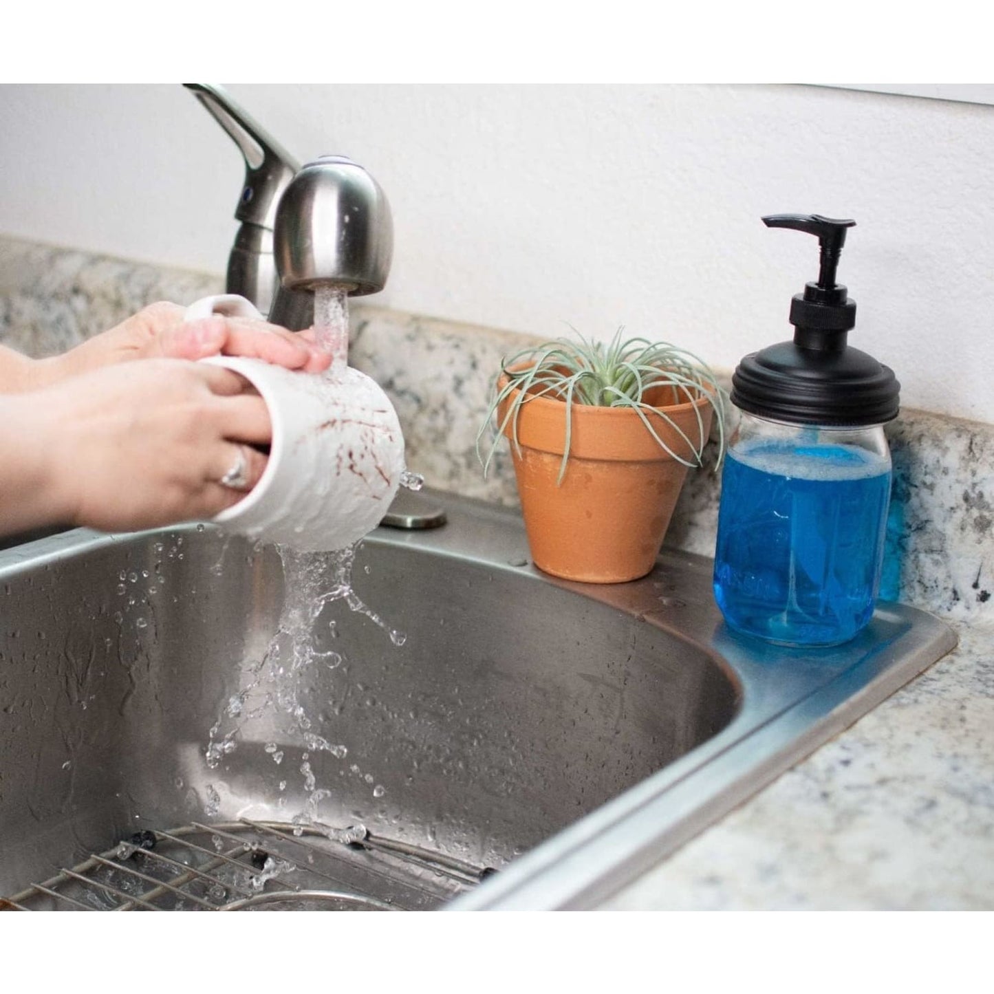 Black pump lid fitted on a clear glass jar filled with blue soap sitting on a kitchen sink countertop next to a small potted plant, ideal for Health & Wellness and Household Helpers collections.