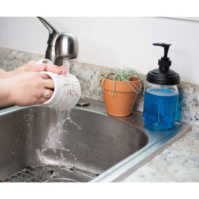 Black pump lid fitted on a clear glass jar filled with blue soap sitting on a kitchen sink countertop next to a small potted plant, ideal for Health & Wellness and Household Helpers collections.