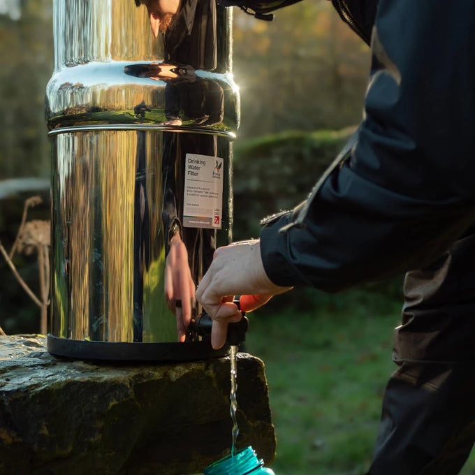 Person filling a container with fresh water from a polished stainless steel gravity water filter outdoors, suitable for camping, emergency supplies, farm, and garden use.