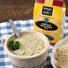 Creamy wild rice soup served in a ceramic bowl with fresh parsley garnish and spoon, accompanied by crackers and Maggie and Mary's handcrafted soup mix packaging, ideal for Kitchen & Food, Dry Goods and Mixes, Soups, USA Made, Best Sellers, and Promo Eligible collections.