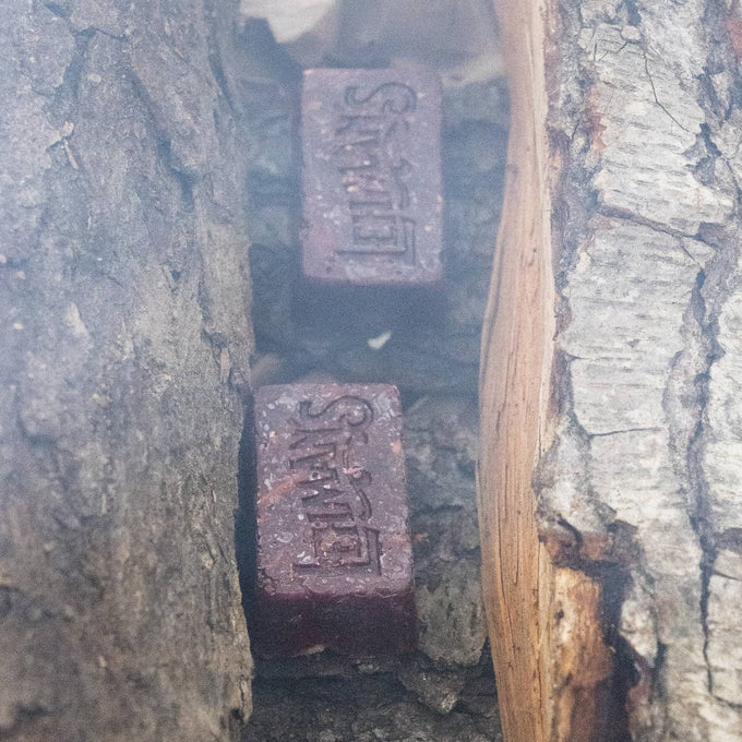 Two handcrafted reddish-brown fire starter blocks embossed with Lehman’s logo, nestled between split logs, made in USA for easy, non-toxic fire building in camping and hearth accessories.