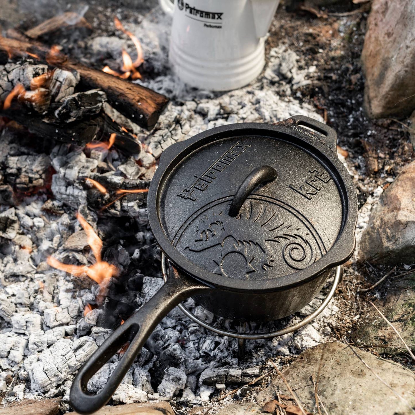 Heavy-duty cast iron saucepan with lid featuring engraved design, cooking over hot embers outdoors, perfect for camping, farm, garden, and cast iron cookware collections.