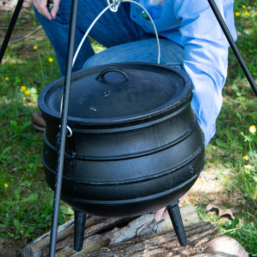 Handcrafted cast iron campfire kettle with rounded shape and three legs, suspended from a tripod over a wood fire, designed for durable outdoor cooking in Farm, Garden & Tool, Camping, Best Sellers, and Promo Eligible collections.