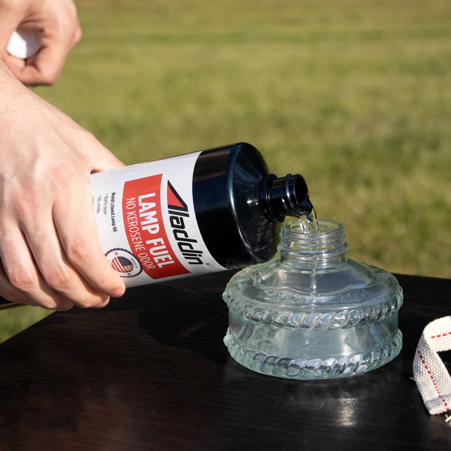 Person pouring clear Aladdin genuine lamp oil from a black 32 oz bottle labeled no kerosene odor into a glass lamp reservoir, set outdoors on a dark table, fitting Lamps & Lighting and Lamp and Lantern Fuel collections.