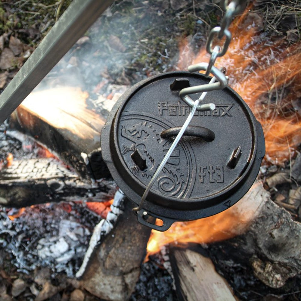 Petromax cast iron Dutch oven with flat base suspended over an open campfire using a metal chain, featuring a detailed embossed lid design, ideal for Kitchen & Food, Cookware, Cast Iron, Best Sellers, Promo Eligible collections.