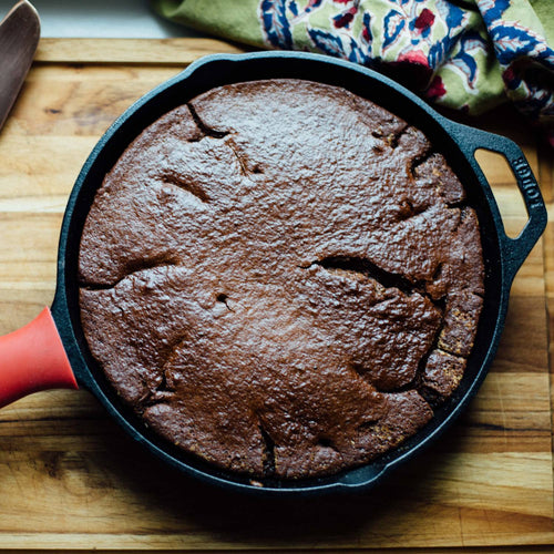 Black cast iron skillet with a freshly baked rich chocolate cake, featuring a red silicone handle cover, shown on a wooden surface with a patterned kitchen towel in Kitchen & Food, Cookware, Cast Iron, Best Sellers, and Promo Eligible collections.