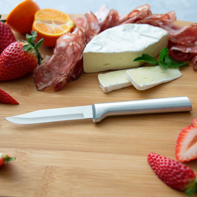 Sharp carbon stainless steel paring knife with a satin-finished cast aluminum handle resting on a wooden board surrounded by sliced strawberries, orange, salami, and creamy cheese, ideal for food prep in USA made and best sellers collections.