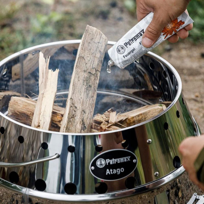 Person pouring liquid fire starter into a stainless steel Petromax Atago Camp Grill and Fire Bowl filled with wood, designed for outdoor cooking and cozy fires in Farm, Garden & Tool and Camping collections.