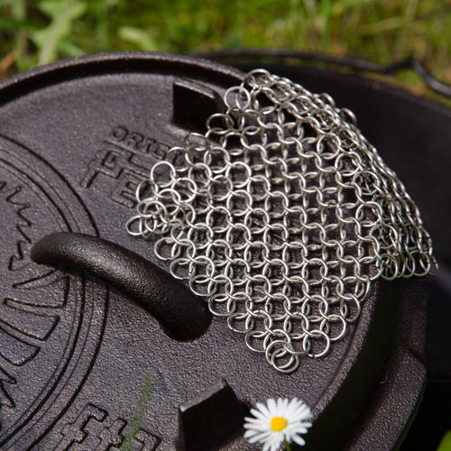 Handcrafted stainless steel chainmail cleaner with interlocking rings resting on a cast iron lid outdoors, designed for tough scrubbing, featured in Home & Decor and Household Cleaning collections.