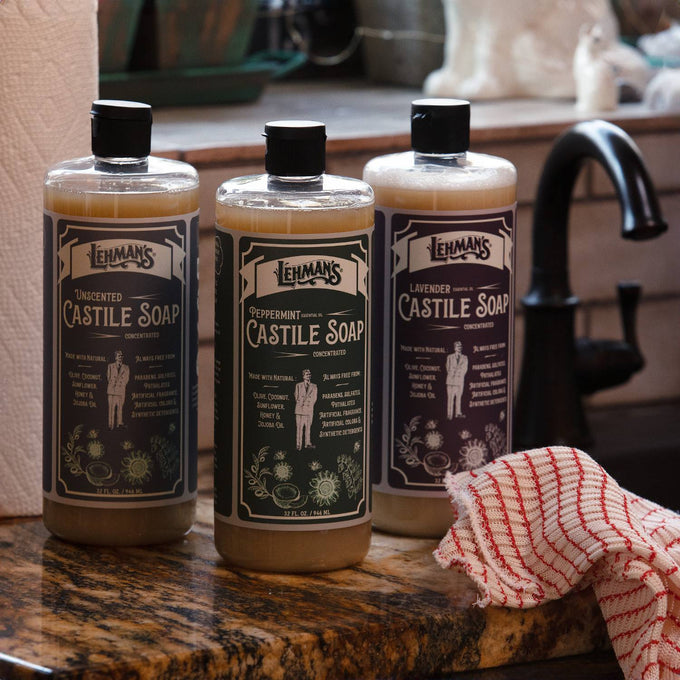Three amber bottles of handcrafted Lehman's multi-use concentrated Castile liquid soap in unscented, peppermint, and lavender variants displayed on a kitchen countertop near a faucet and dish towel, highlighting natural USA made products in Health & Wellness, Grooming and Personal Hygiene, and Soaps collections.