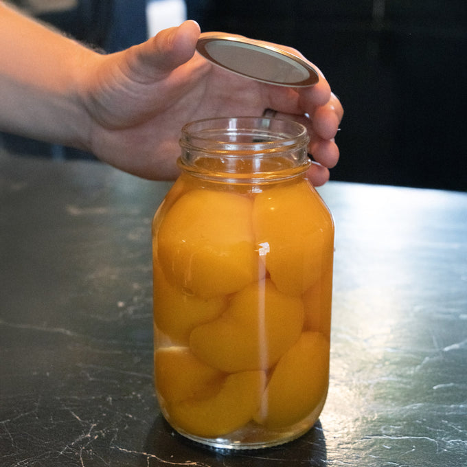 Hand holding a silver-toned regular-mouth canning jar lid with BPA-free sealing compound above a glass jar filled with preserved peaches, suitable for Kitchen & Food and Food Preservation collections.