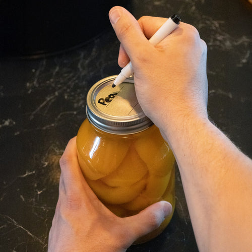 Hands holding a glass jar filled with preserved peaches, sealing it with a regular-mouth canning lid and band, while labeling the lid with a marker, set on a dark kitchen counter.