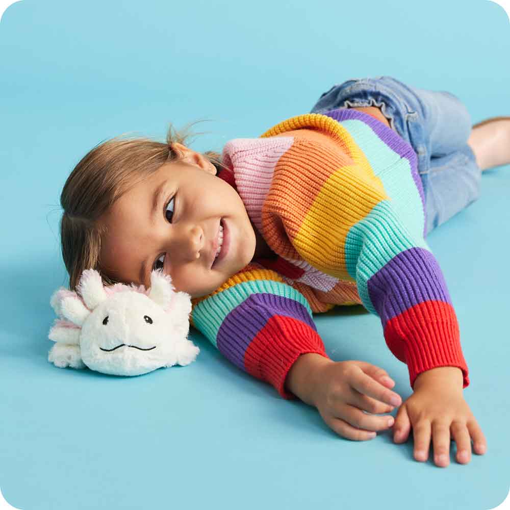 Child smiling and lying on blue floor cuddling a soft, microwaveable white plush axolotl scented with French lavender, ideal for Health & Wellness, Children's Toys, and Indoor Play collections.