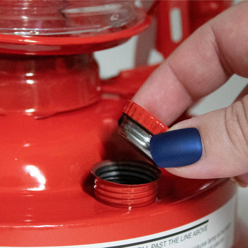 Hand removing a small red fuel cap from a bright red handcrafted lantern with weather-resistant finish, ideal for Farm, Garden & Tool, Camping, and Emergency Supplies collections.