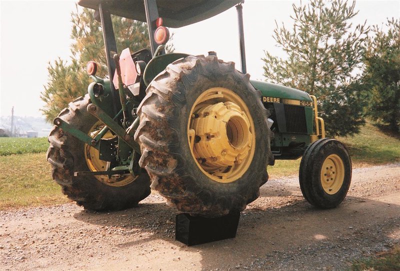 Green and yellow John Deere tractor positioned on a rural dirt path with a sturdy black support block beneath the rear tire, surrounded by pine trees and open farmland.