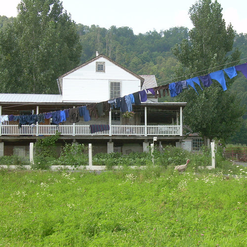 Long clothesline supported by trees and porch posts holding dark blue and purple garments drying outdoors near a traditional farmhouse in a rural green landscape, showcasing handmade Amish quality for home and decor use.