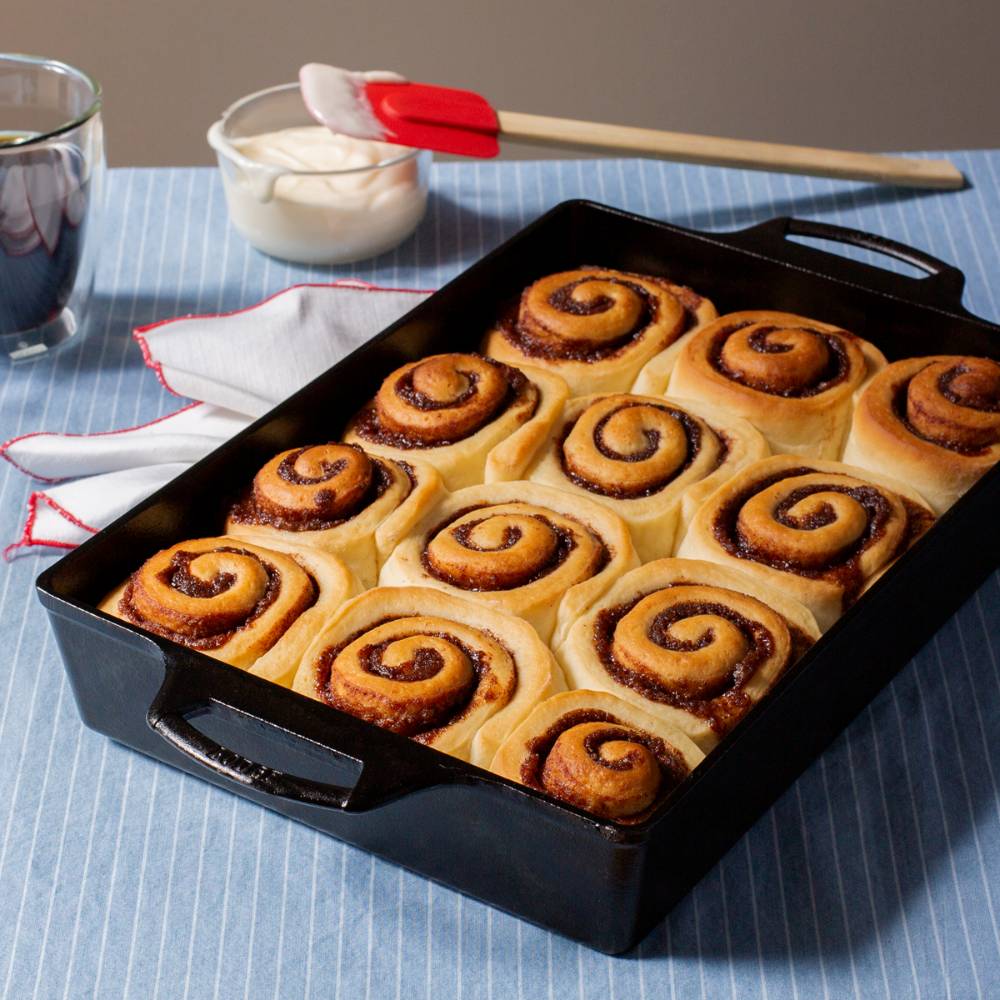 Black cast iron casserole pan with dual handles freshly baked cinnamon rolls arranged inside on a blue striped cloth with a red silicone brush and bowl of icing nearby, ideal for Kitchen & Food, Cookware, Cast Iron, USA Made, Best Sellers, and Promo Eligible collections.